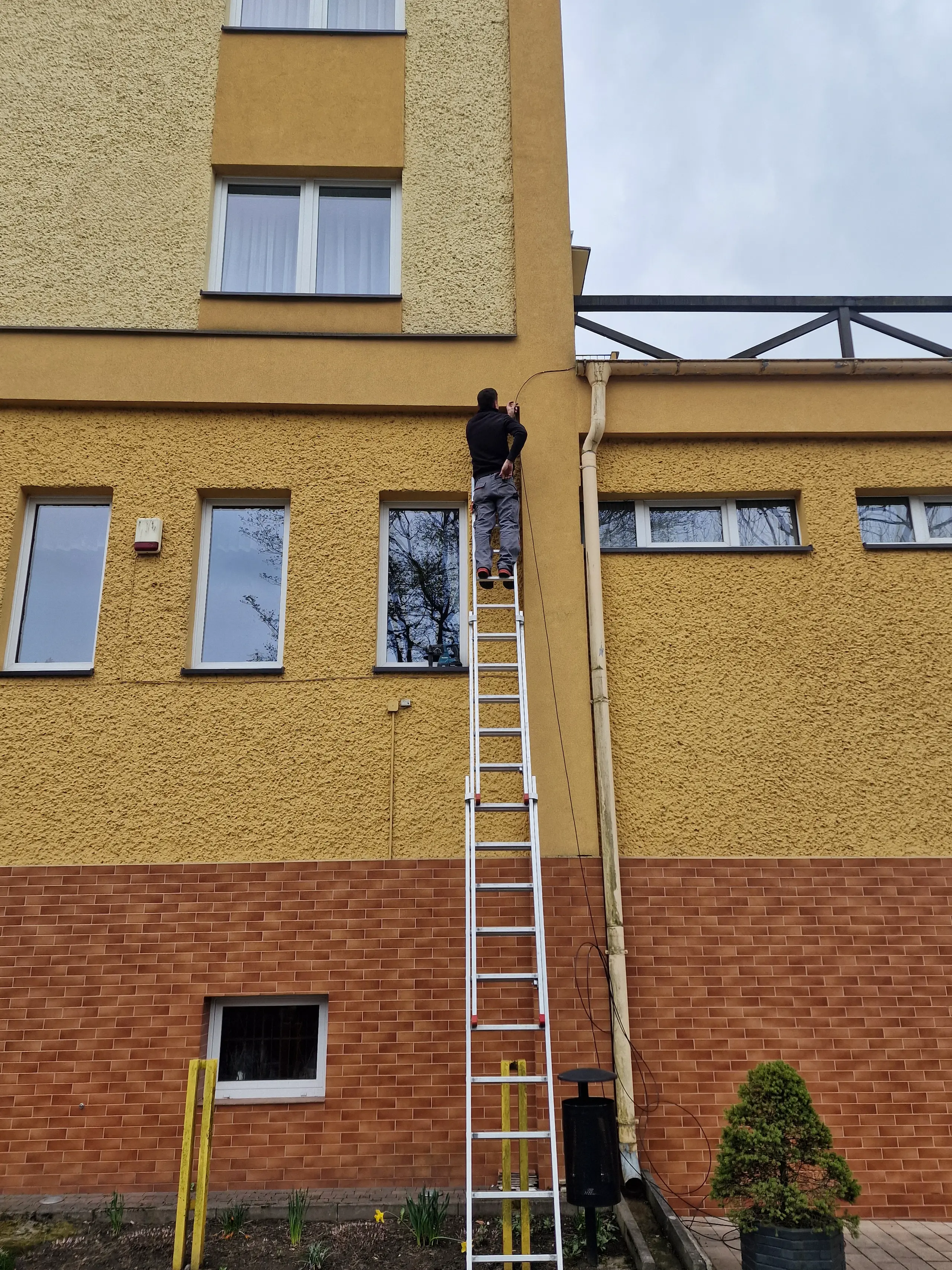 Montaż instalacji elektrycznej na budynku, pracownik na drabinie przy wykonywaniu prac
