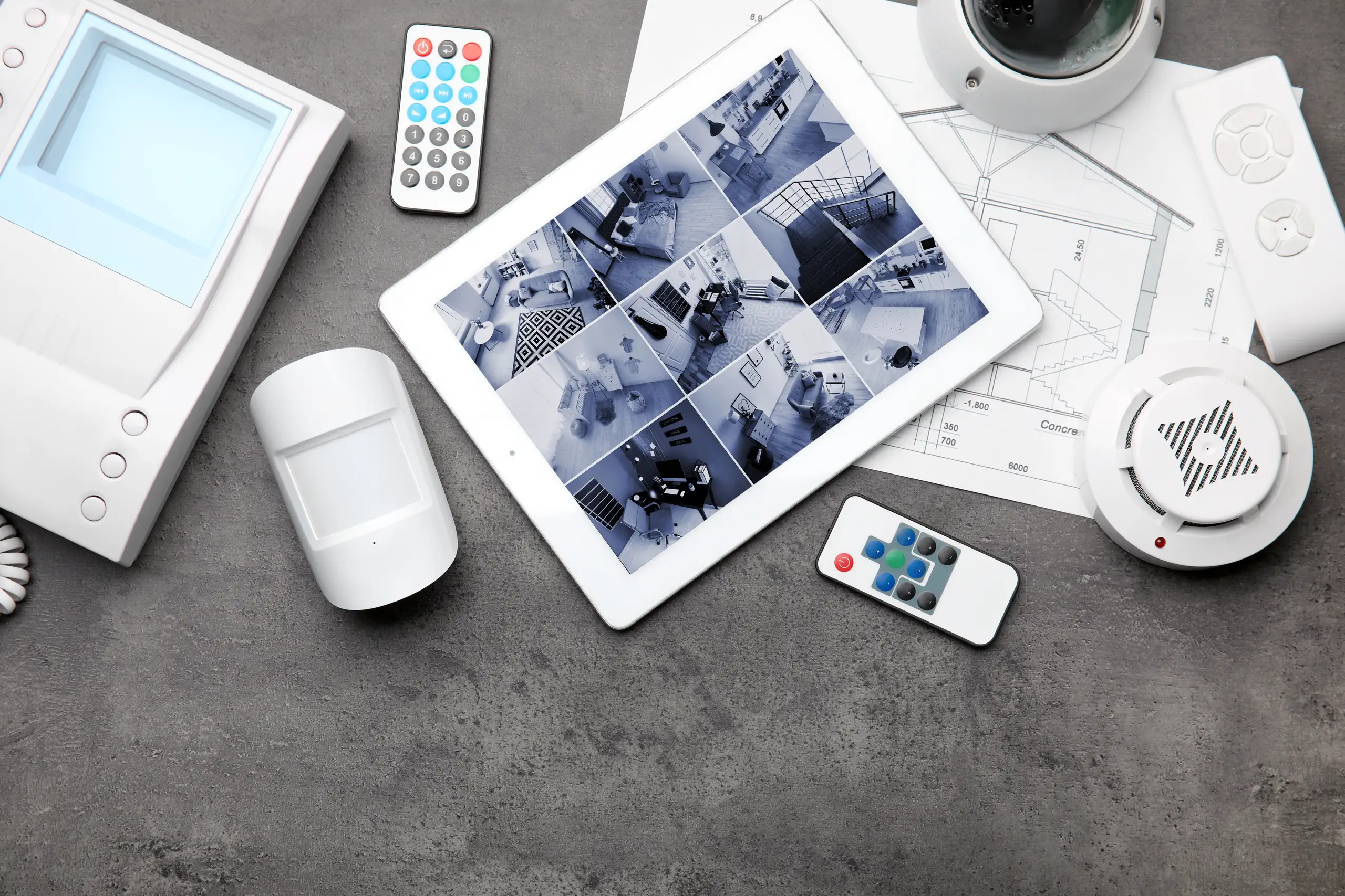 Tablet z widokiem z kamer monitorujących, obok pilota i czujników alarmowych